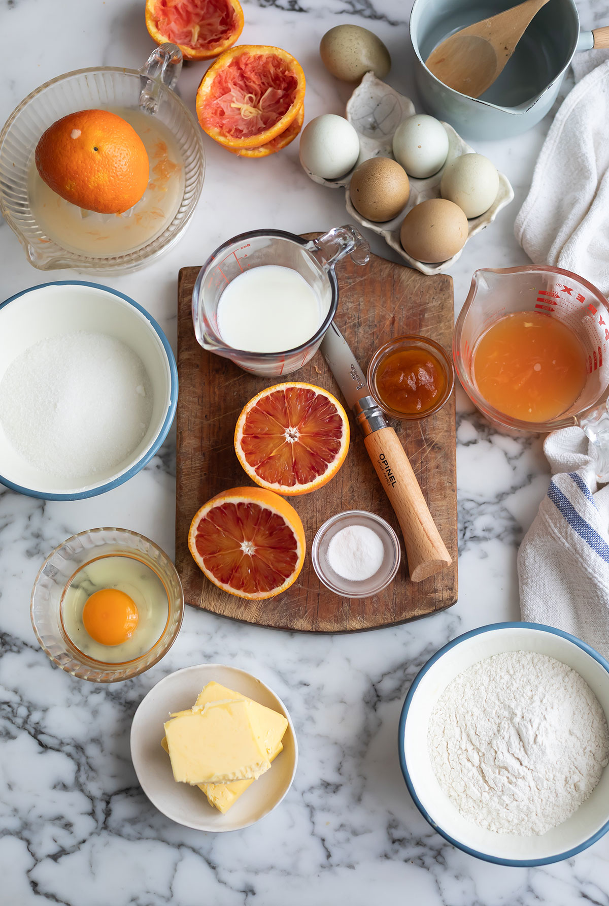 Ingredients for the best malva pudding recipe - a classic South African dessert • Drizzle and Dip Top-down view of Malva pudding ingredients on a marble counter: orange halves, eggs, milk, butter, sugar, flour, and a wooden knife on a cutting board.