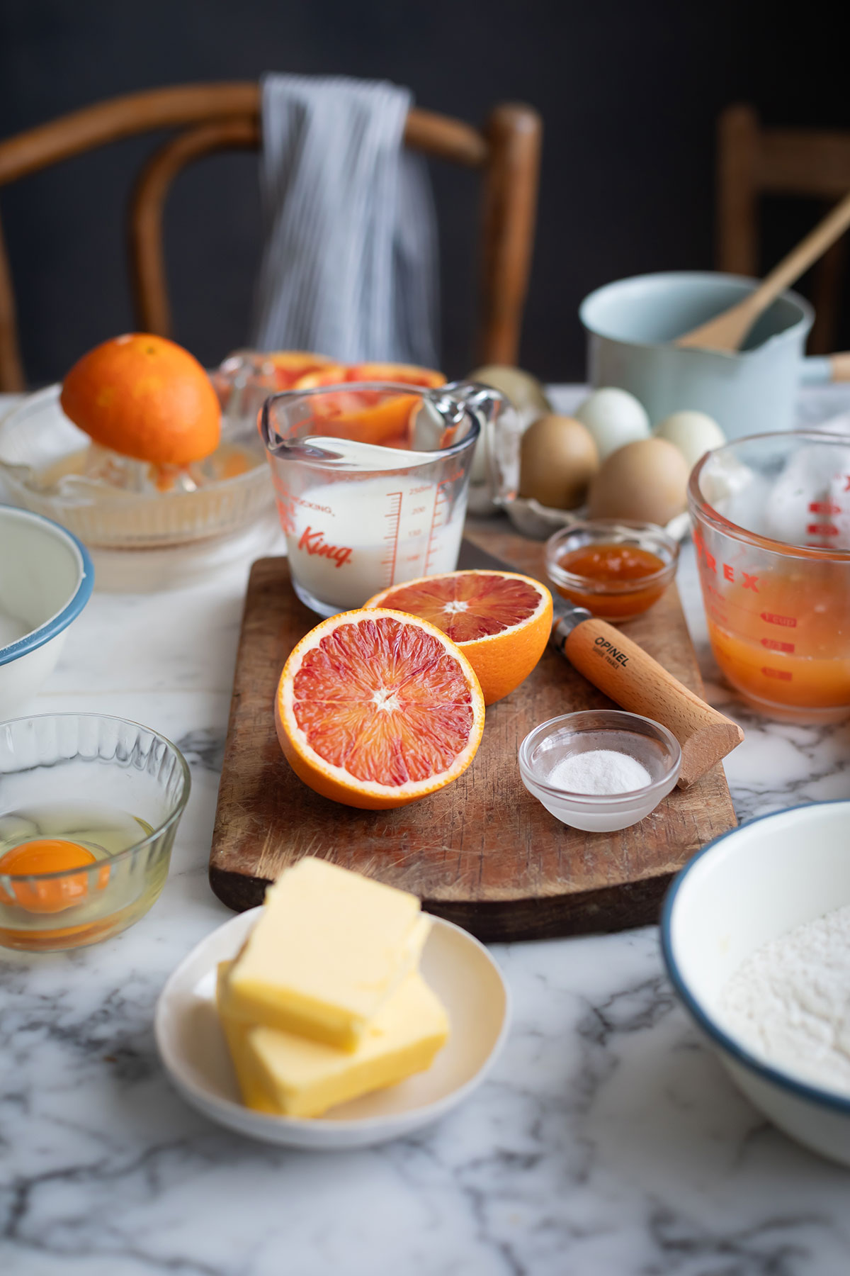 Malva pudding with orange recipe • Drizzle and Dip Half orange on a wooden board among baking ingredients like butter, eggs, flour, and measuring cups on a marble countertop for a malva pudding recipe.