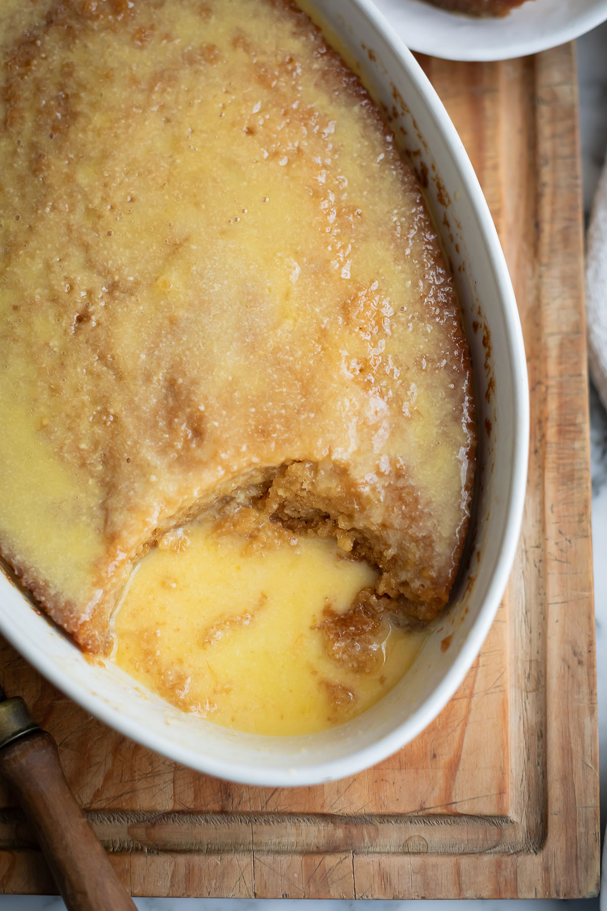 The best South African malva pudding recipe • Drizzle and Dip Golden Malva pudding topped custard in a white oval dish on a wooden board, with a scoop taken out.
