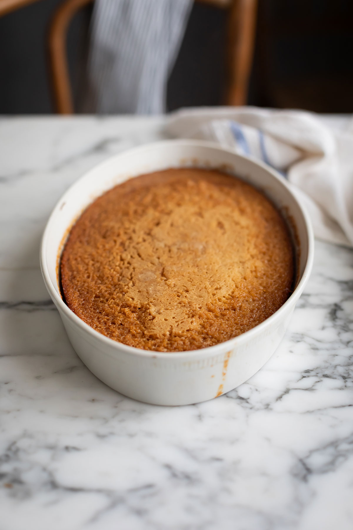 aked Mlava pudding without sauce • Drizzle and Dip Golden brown malva pudding cooling in a white round baking dish on a marble countertop, with a striped cloth in the background.