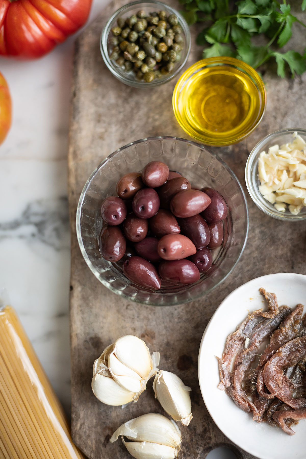 Olives for spaghetti puttanesca pasta sace recipe • Drizzle and Dip Assortment of cooking ingredients on a wooden board: olives in a glass bowl, capers, olive oil, garlic, parsley, parmesan shavings, spaghetti, a tomato, and sliced meat on a plate.