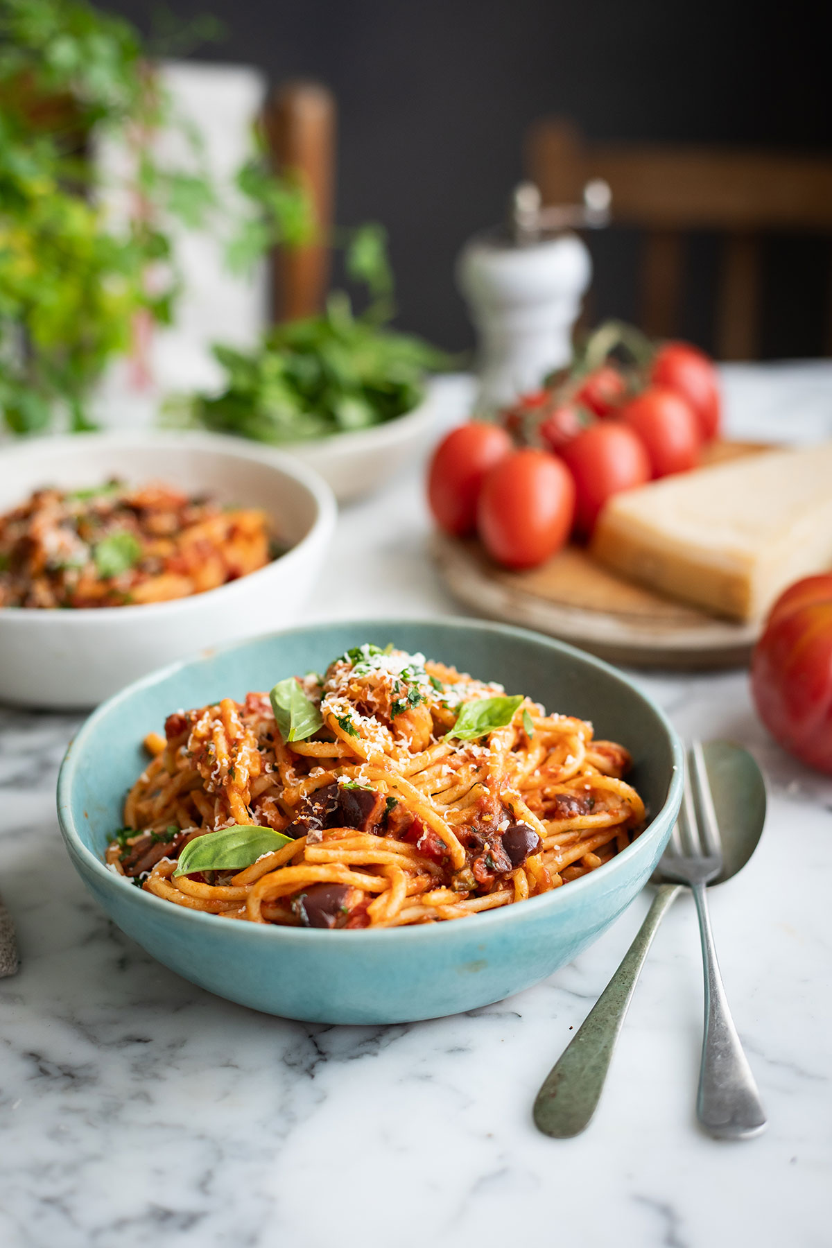 Spaghetti puttanesca with tomatoes, olives, capers and anchovies recipes • Drizzle and Dip Blue bowl of spaghetti puttanesca with tomato sauce, cheese, and fresh basil on a marble countertop.