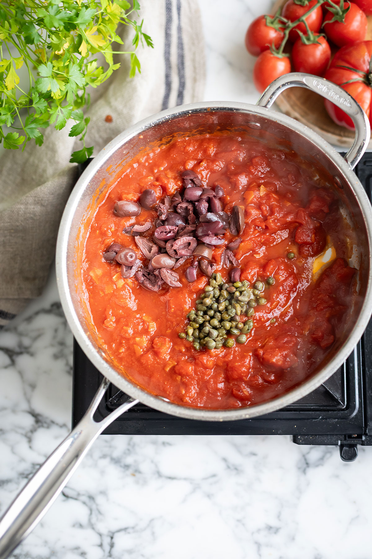 How to make spaghetti puttanesca, a classic Neopilitan pasta recipe • Drizzle and Dip Tomato sauce simmering in a pot on the stove, topped with sliced olives and green capers, with fresh herbs and tomatoes nearby.